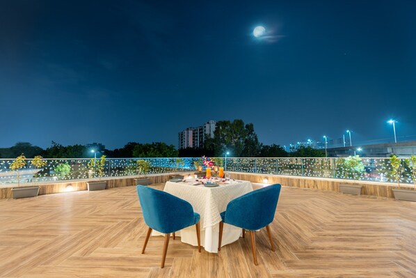 An outdoor dining area with a table set for two, surrounded by blue chairs, under a night sky with a visible moon.