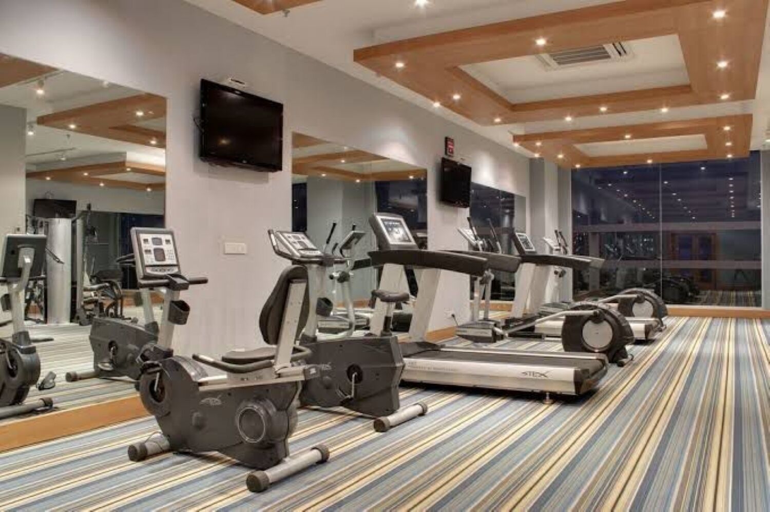 Un gimnasio con cintas de correr, elípticas y bicicletas estáticas, espejos grandes en las paredes y una alfombra estampada.