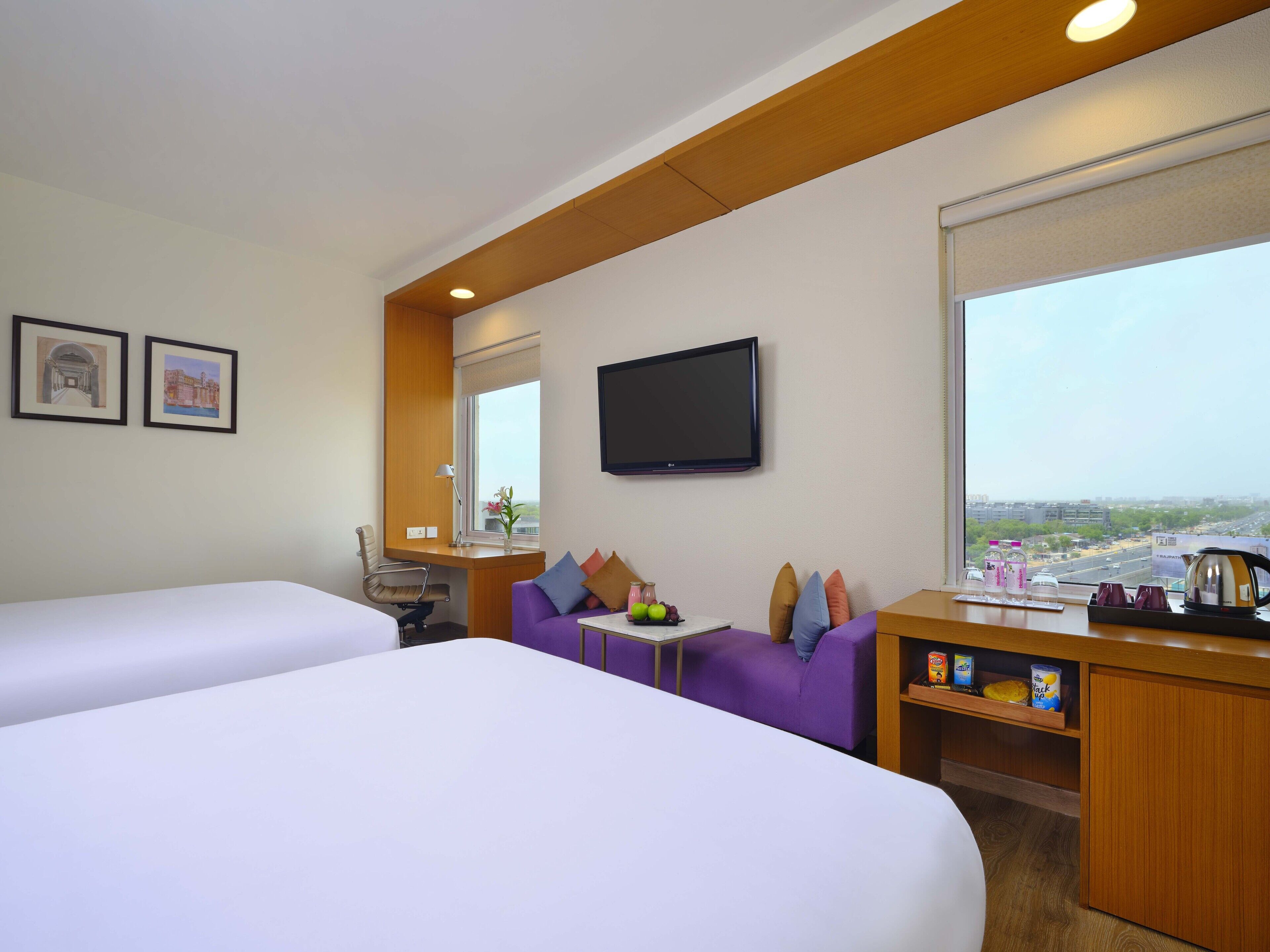 A hotel room with two beds, a sofa, a TV, a window with a city view, and a small table with a lamp and a vase of flowers.