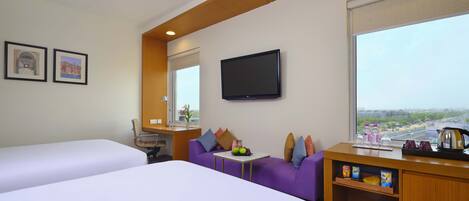 Habitación de hotel con dos camas, un sofá, un televisor, una ventana con vista a la ciudad y una mesita de noche con una lámpara y un florero.