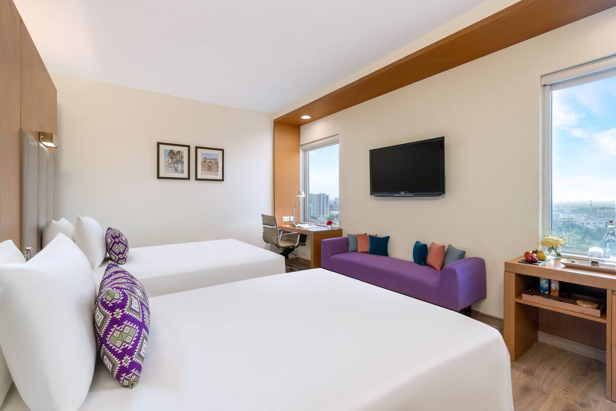 Ein Hotelzimmer mit zwei Betten, einem Schlafsofa, einem Schreibtisch mit Fernseher und einem Fenster mit Blick auf die Stadt.