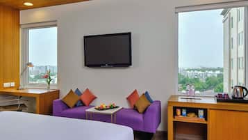 A hotel room with a bed, a purple sofa, a small table with snacks, a TV on the wall, and a large window with a city view.