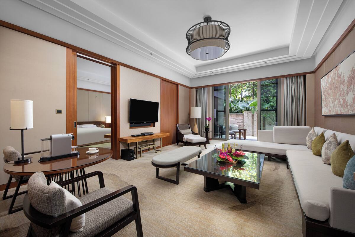 A modern hotel room with a large sofa, a dining table, a TV, and a view of the outdoors.