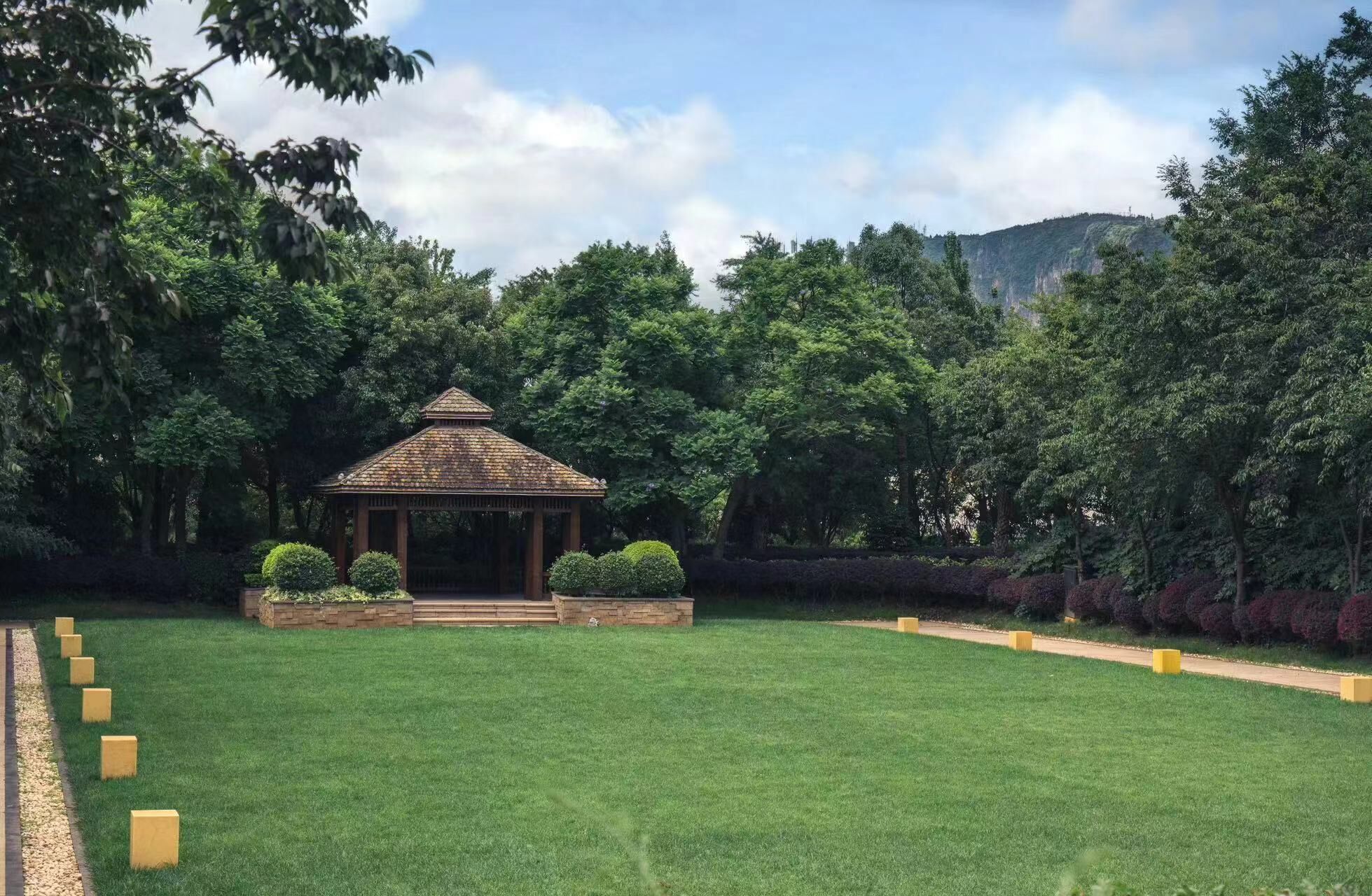 A gazebo in a park with a well-maintained lawn, surrounded by dense trees and a mountain in the background.