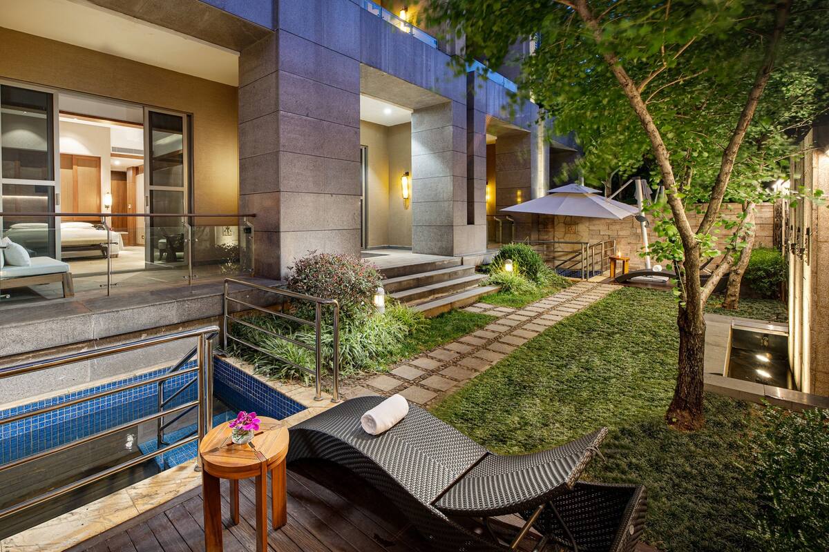 A modern building with a glass entrance, a pool area with a hot tub, and a small garden with a tree and outdoor furniture.