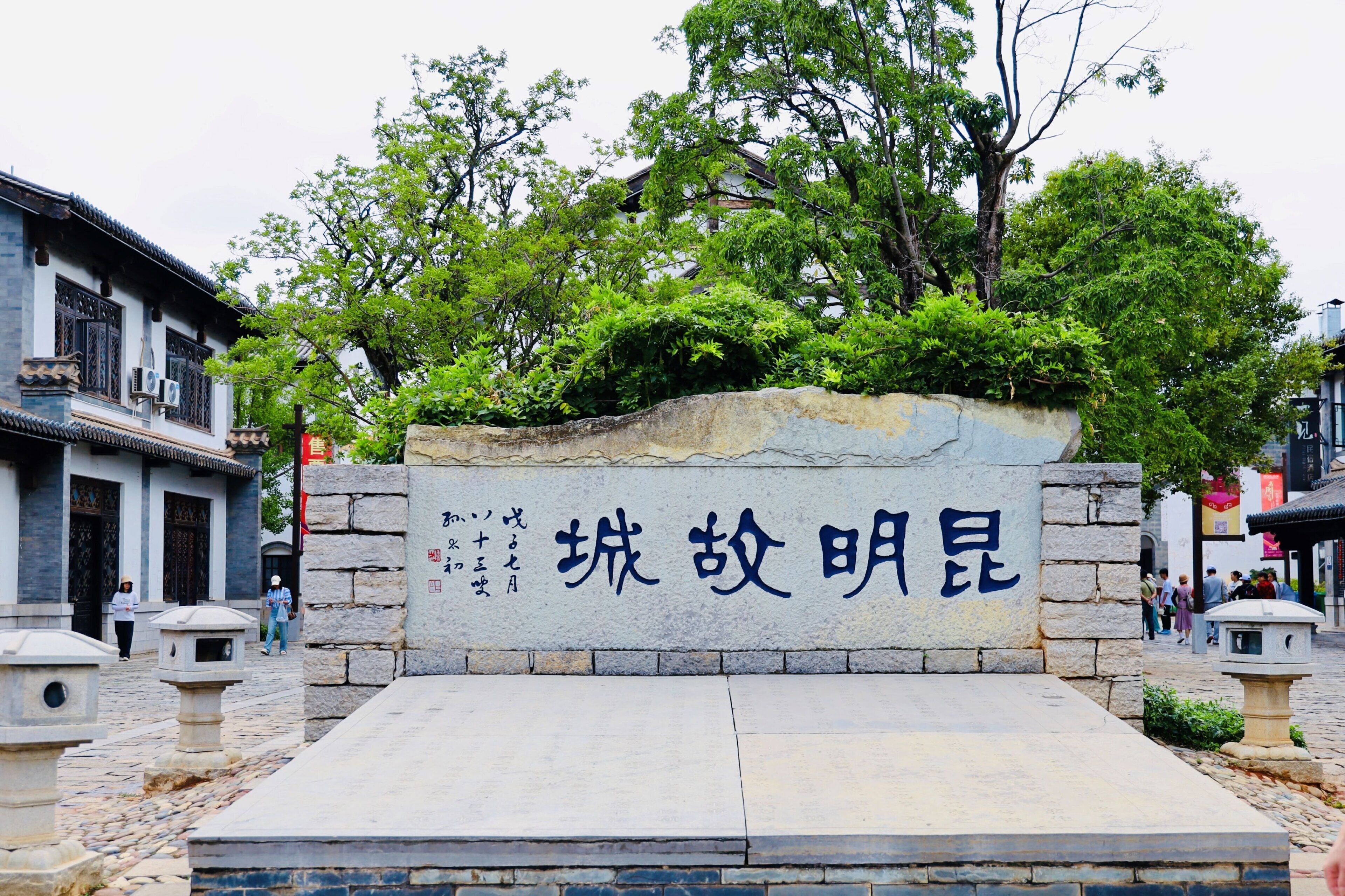 A stone wall with Chinese characters, a traditional building, and a tree-lined street.