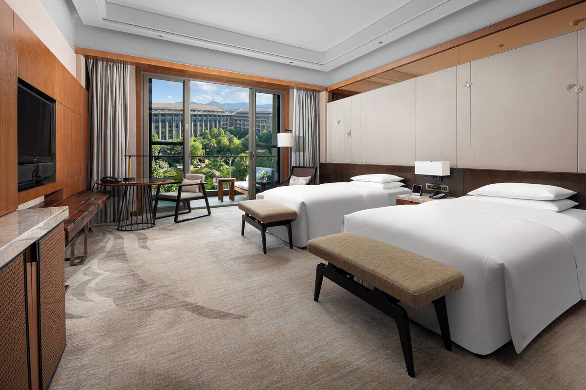 A hotel room with two beds, a desk, a TV, and a view of a cityscape.