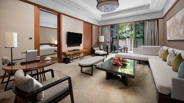 A modern hotel room with a sofa, a coffee table with flowers, a TV, and a desk with a lamp.