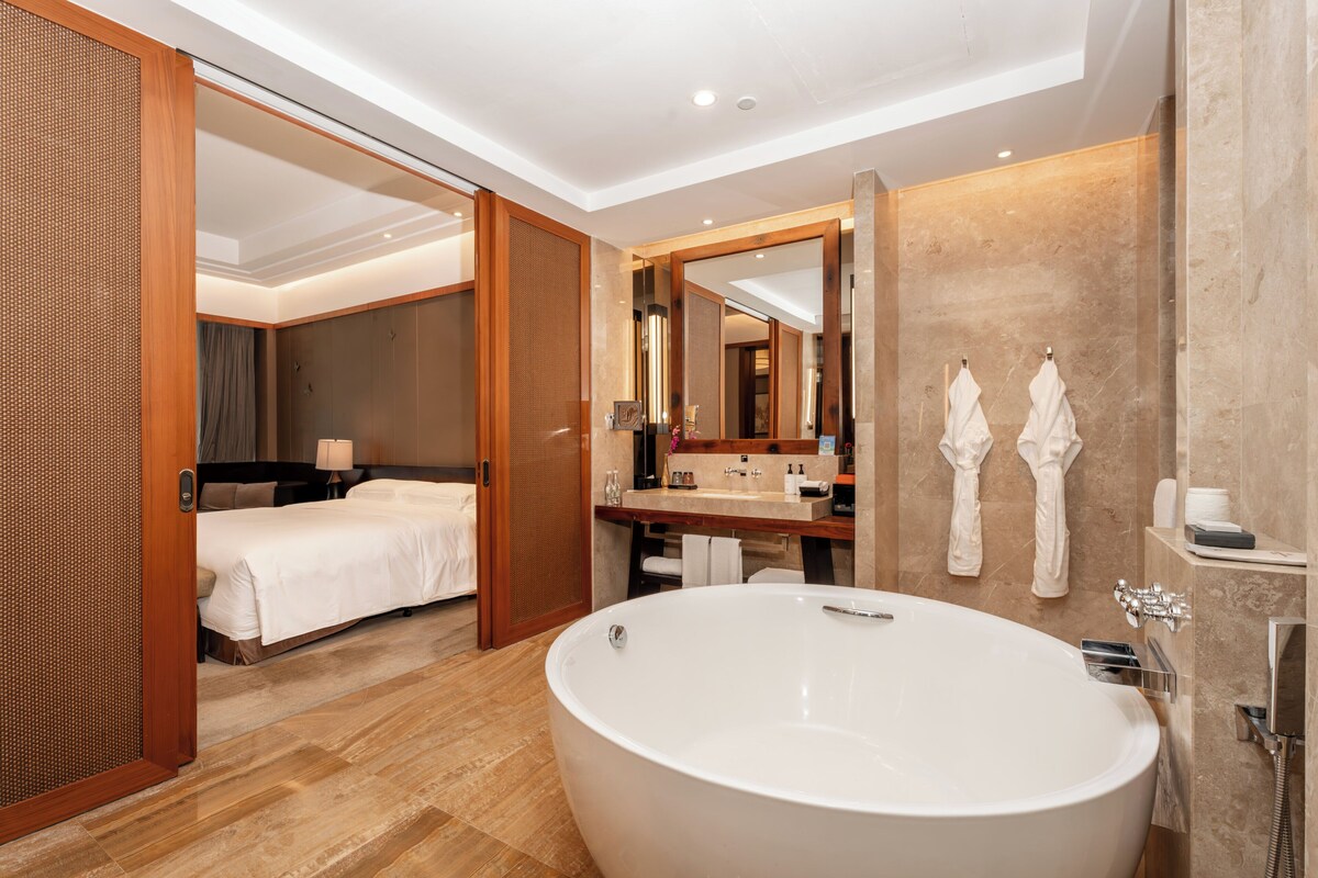 Una habitación de hotel moderna con una bañera grande, un dormitorio independiente, un baño con espejo y toallas, y una puerta de madera.