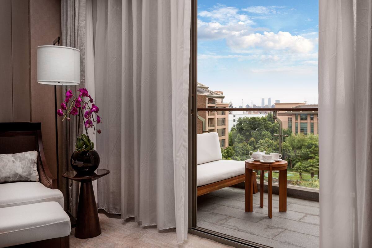 A hotel room with a bed, a small table with a vase of flowers, a chair, and a balcony with a view of buildings and greenery.