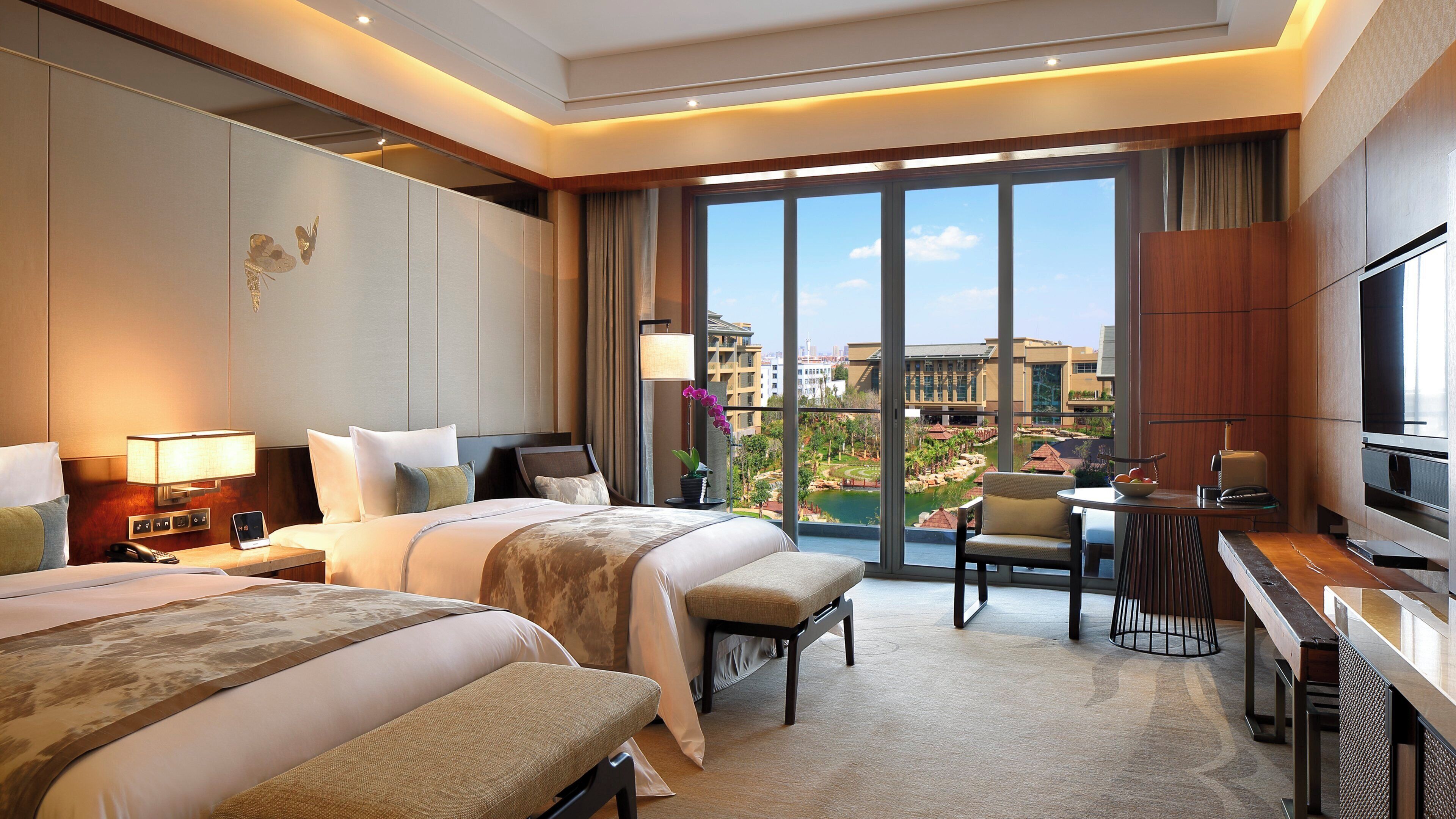 Habitación de hotel con dos camas, un escritorio, una silla, un televisor y un ventanal con vistas a edificios y una masa de agua.