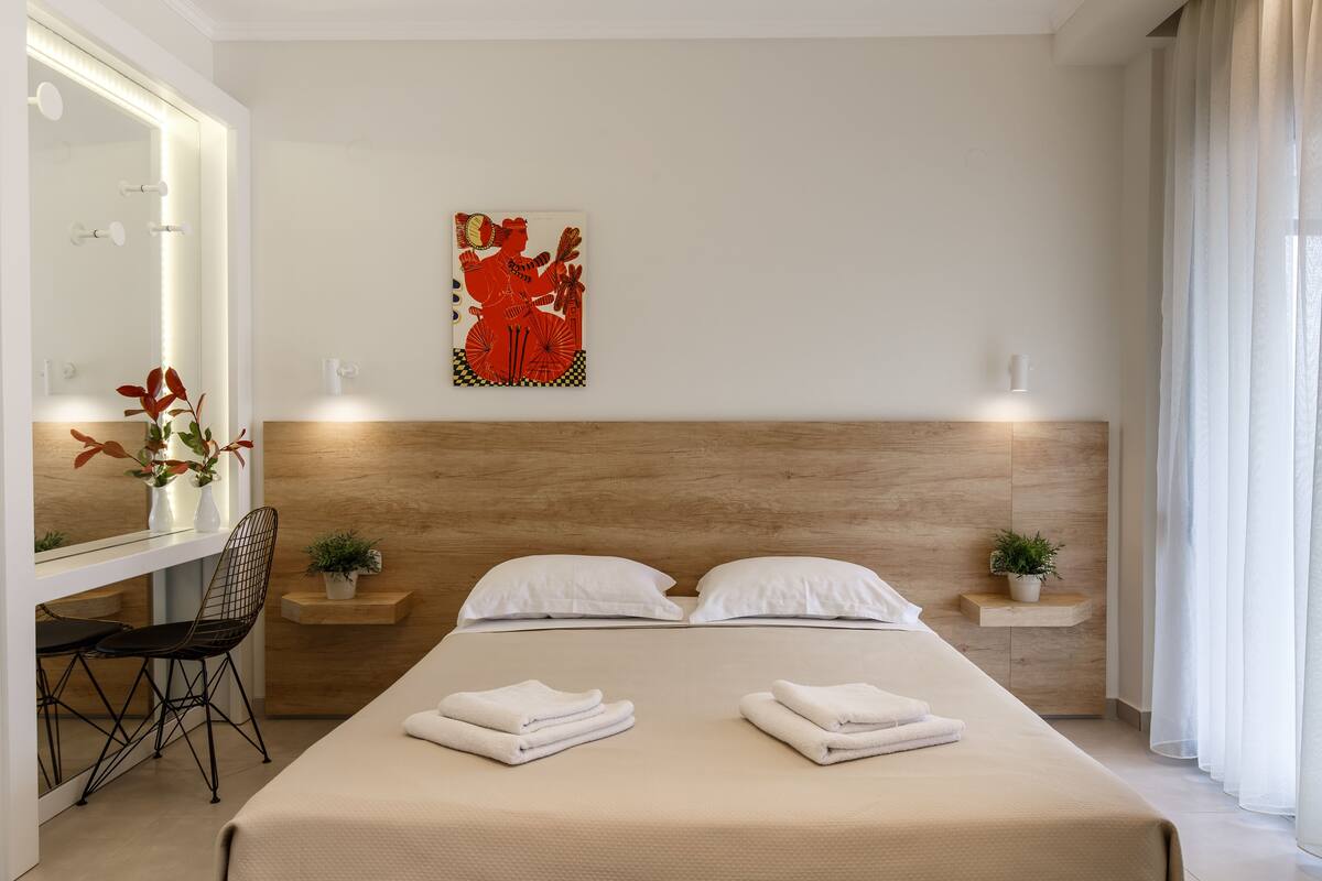 A modern hotel room with a wooden headboard, a large bed with white linens, a desk with a chair, a mirror, and a wall-mounted artwork.