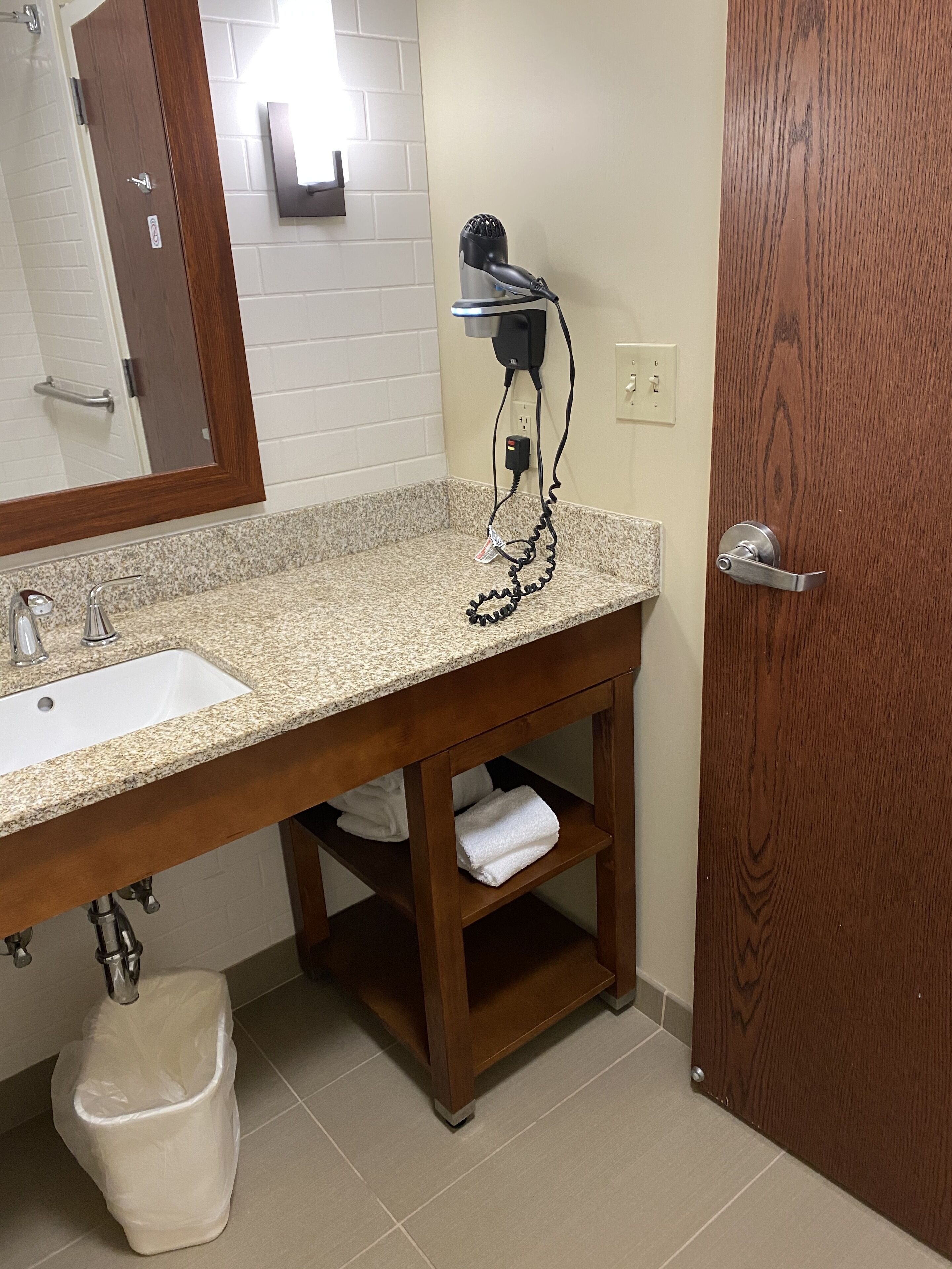 A bathroom with a granite countertop sink, a mirror, a hairdryer on a wall-mounted stand, a towel rack, and a wooden door.