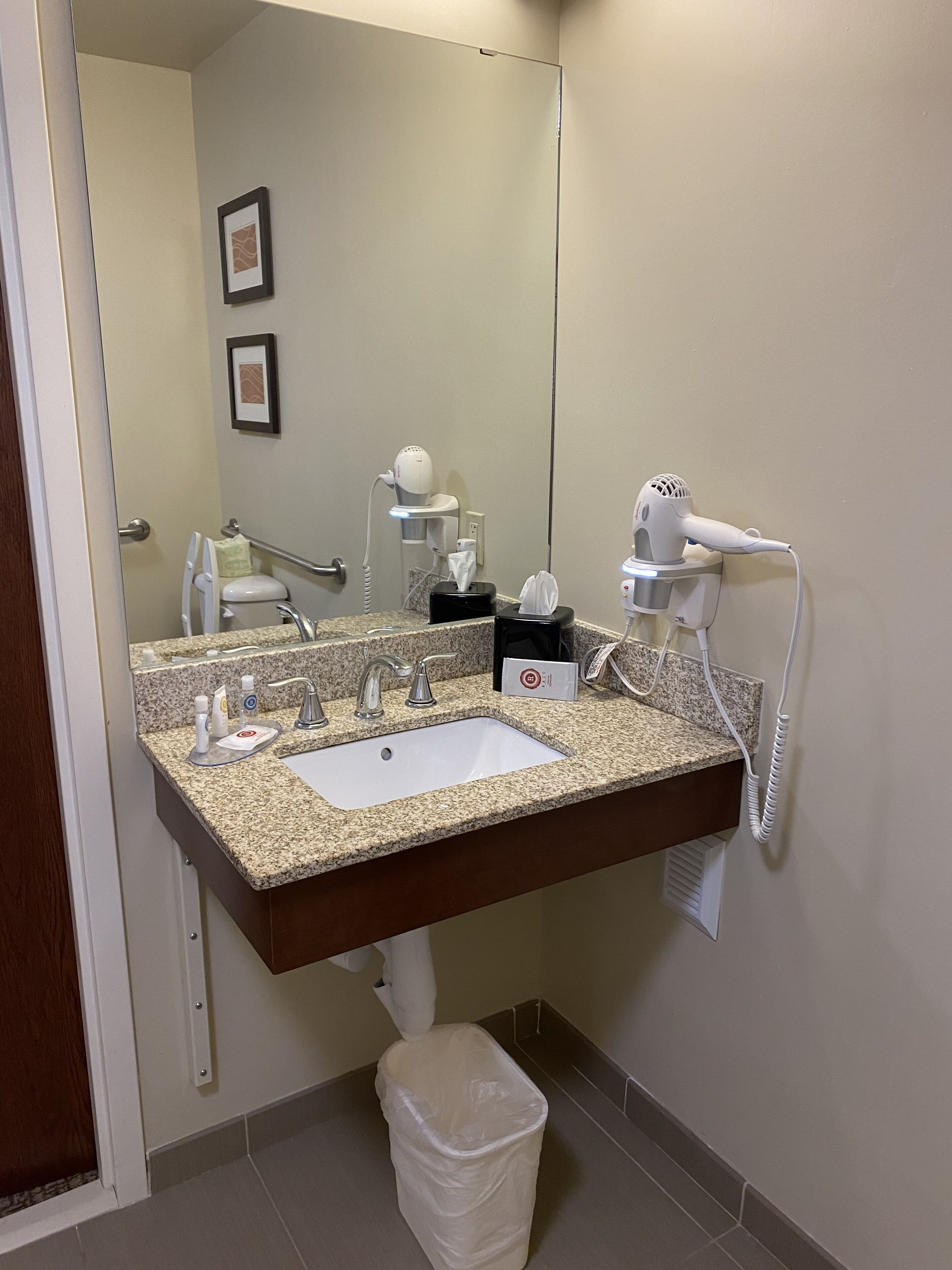 A bathroom with a granite countertop sink, a hairdryer, a mirror, and a trash bin.