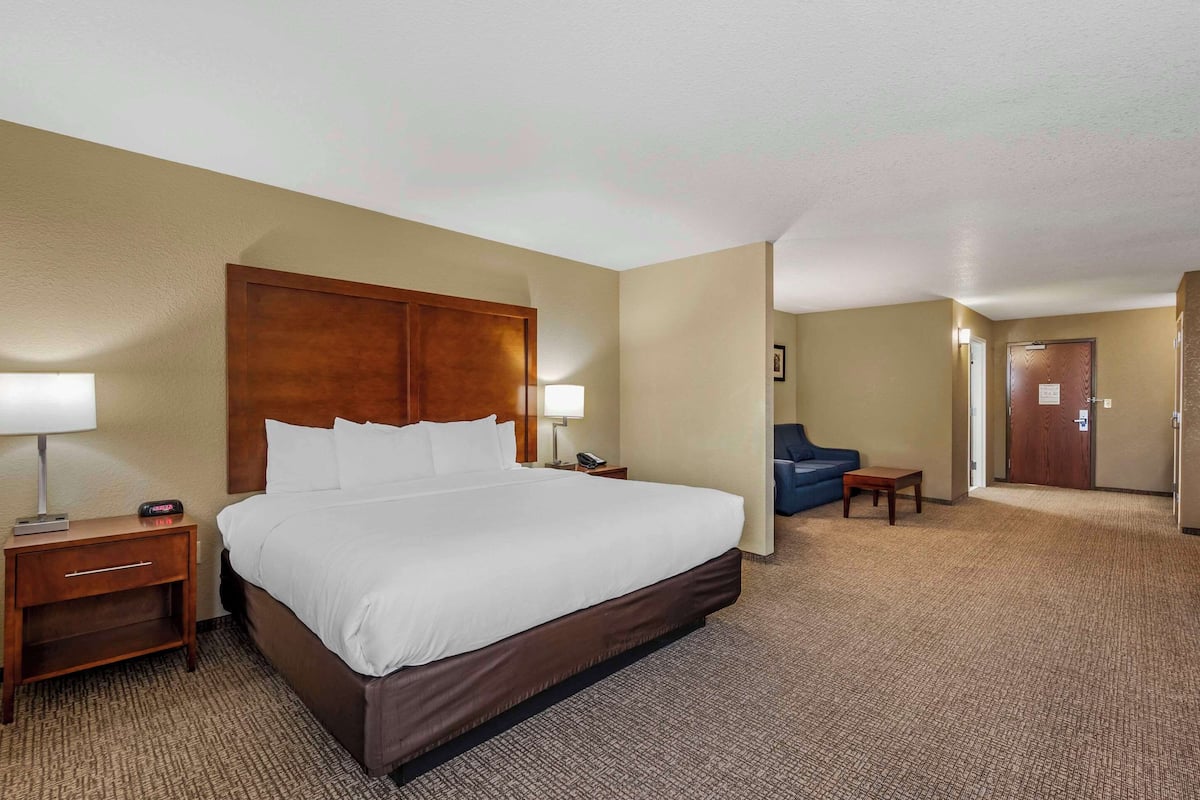 A hotel room with a large bed, a nightstand with a lamp, a small table, and a blue chair.