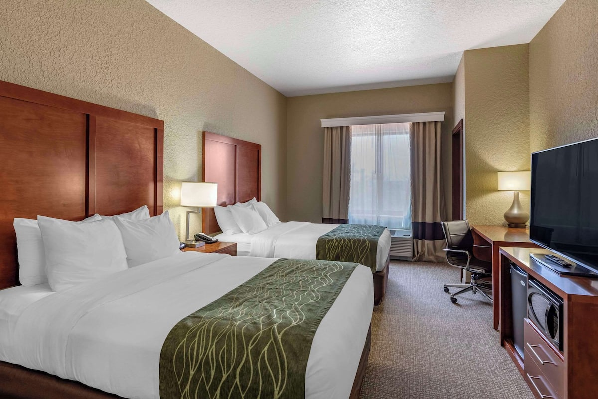 A hotel room with two beds, a desk with a TV, a chair, a lamp, and a window with curtains.