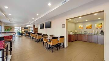 A spacious restaurant with multiple dining tables and chairs, a kitchen area with a buffet, and a large window providing natural light.