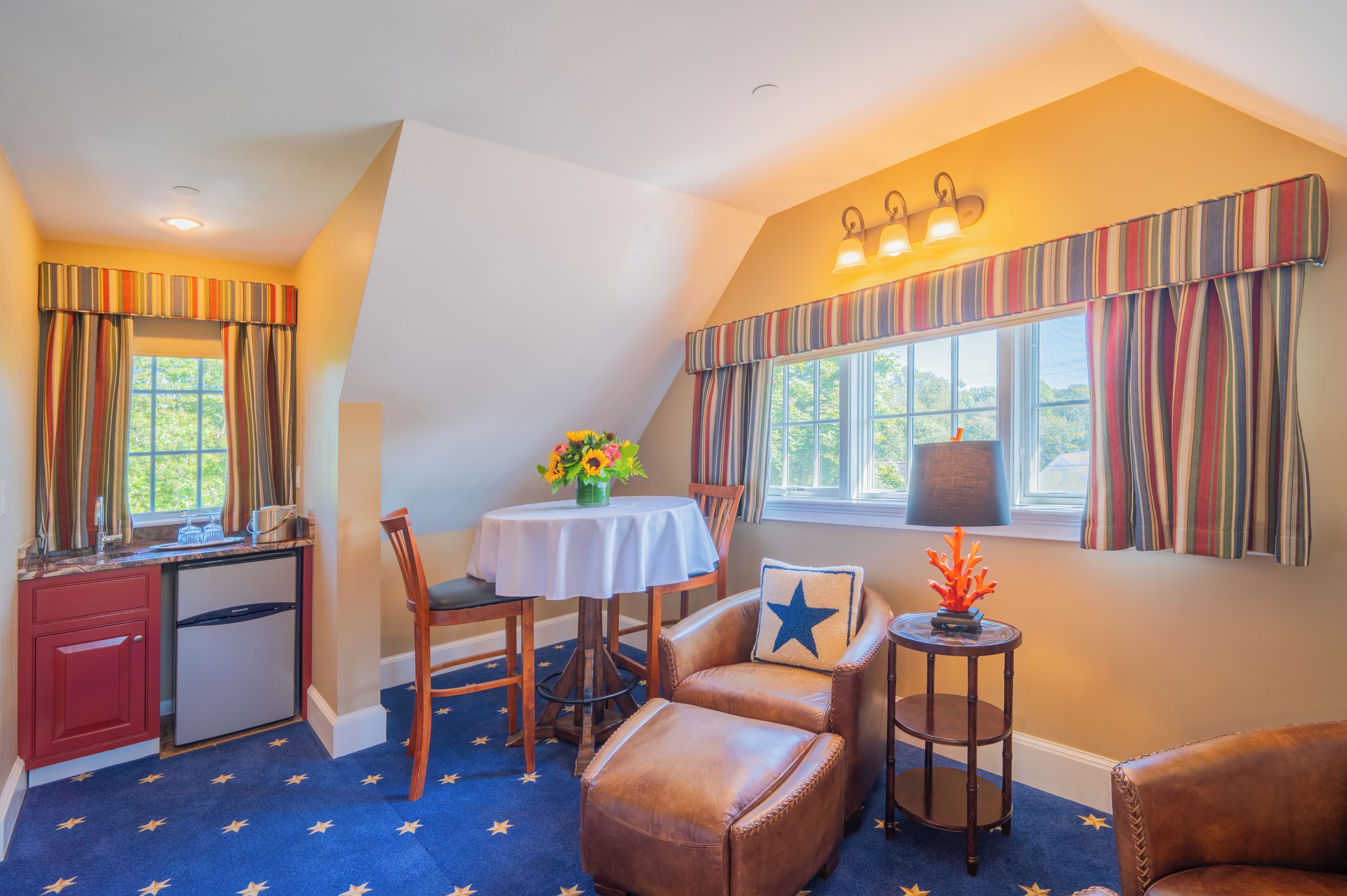 A room with a dining table, chairs, a small side table with a lamp, a decorative pillow, and a window with curtains.