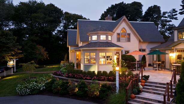 A two-story house with a well-lit front porch, a staircase leading to the entrance, and a garden with various plants and flowers. - Winstead Inn (Harwich)