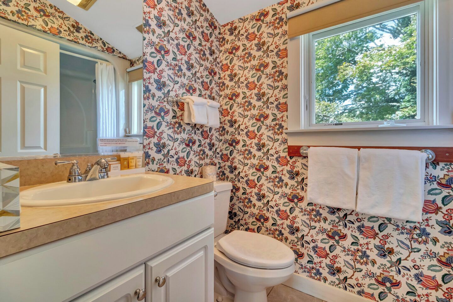 A bathroom with floral wallpaper, a sink, a toilet, and a window with a view of trees.