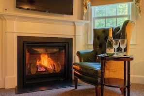 A fireplace with a fire, a leather armchair, a table with a bottle and glasses, and a television mounted above the fireplace.