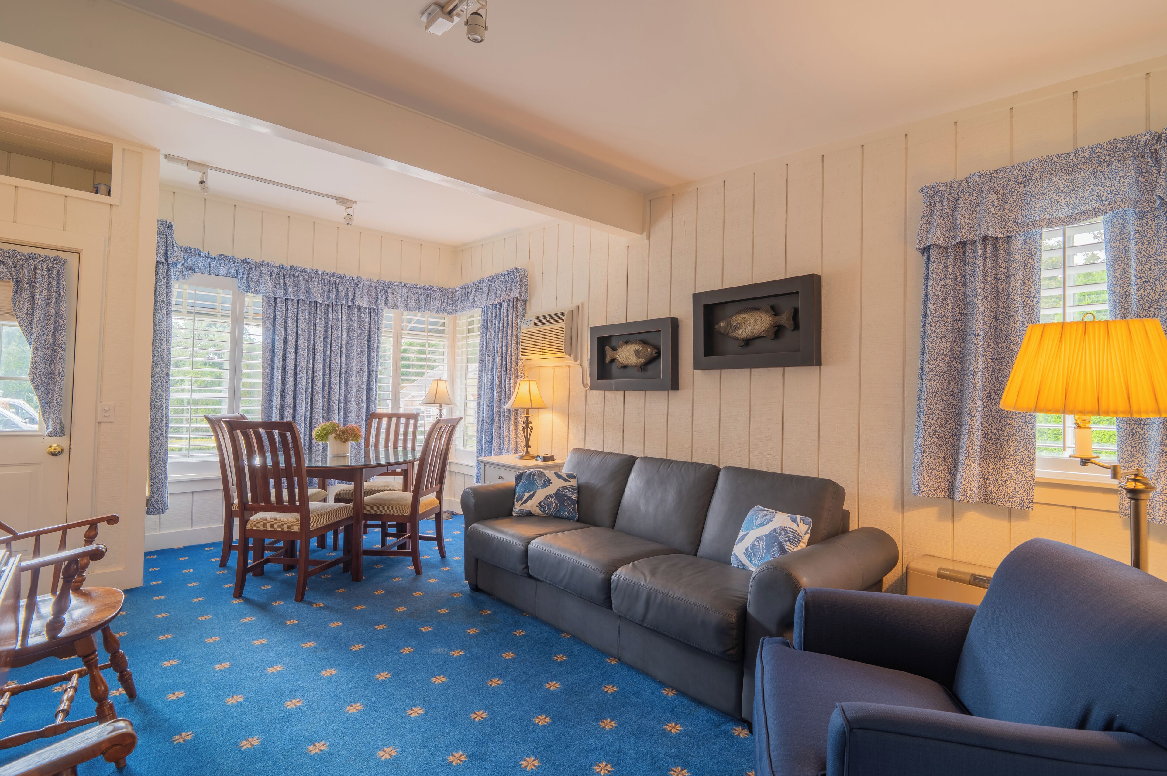 A room with a blue patterned carpet, a leather sofa, wooden chairs, a dining table, and framed fish pictures on the wall.
