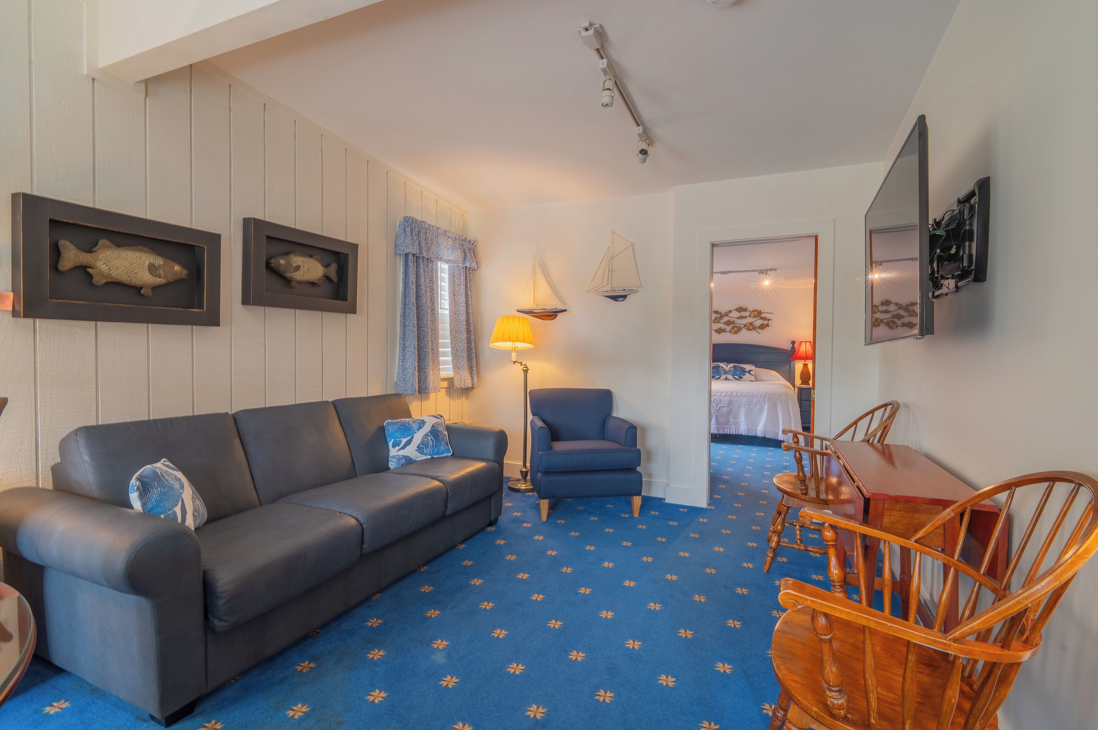 A room with a blue patterned carpet, a dark leather sofa, a wooden dining table with chairs, and framed fish on the wall.
