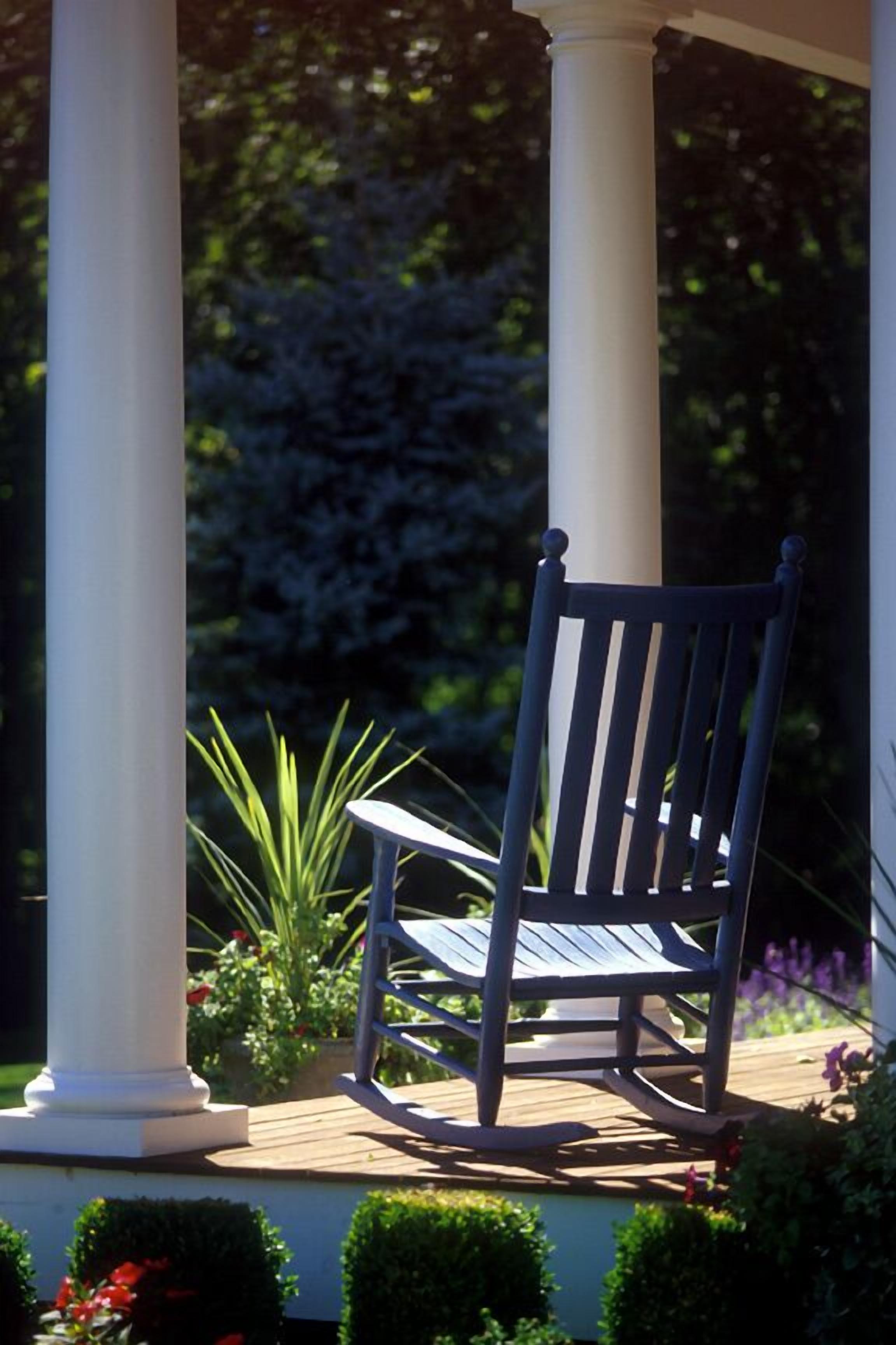 A wooden rocking chair on a wooden deck with white columns, surrounded by greenery and flowers.