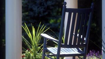 A wooden rocking chair on a wooden deck with white columns, surrounded by greenery and flowers.