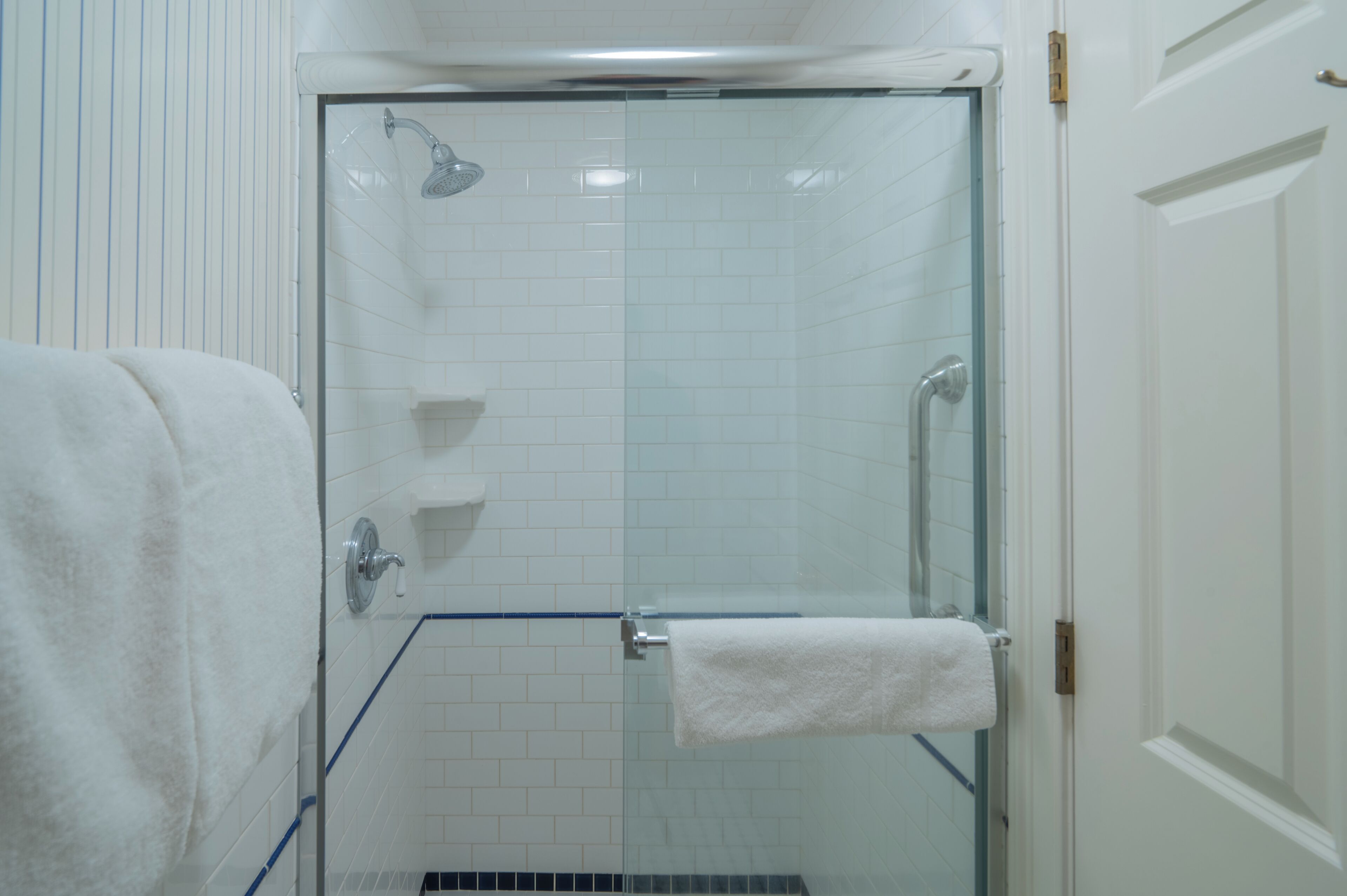 Ducha con cerramiento de vidrio, pared con azulejos blancos, una toalla blanca en la pared y una puerta blanca.