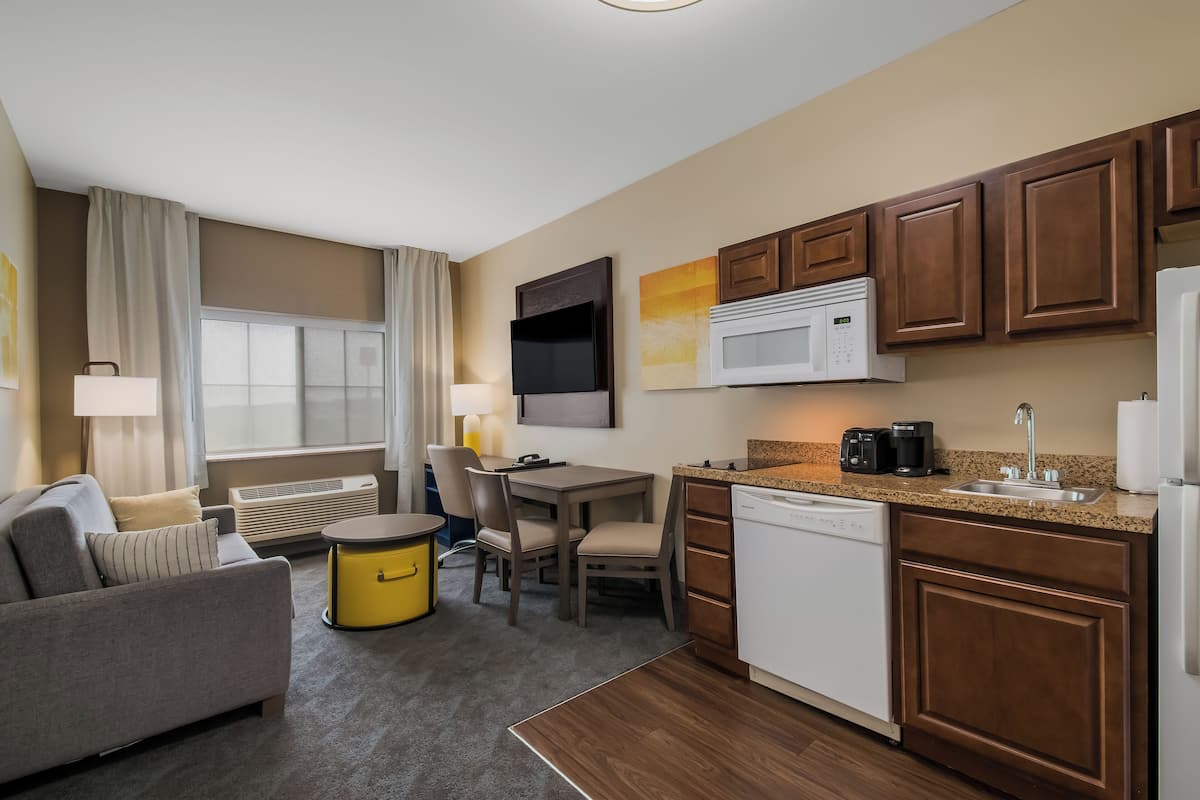A compact hotel room with a sofa, dining table, chairs, a small kitchen with a microwave, coffee maker, and a dishwasher.