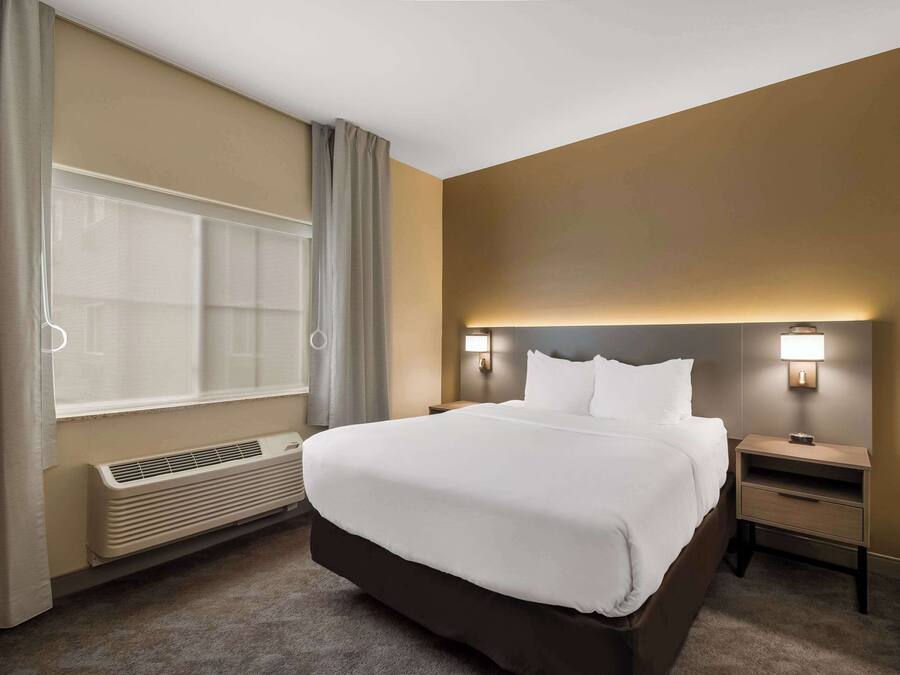 Une chambre d’hôtel avec un grand lit, une table de chevet, une commode, une fenêtre avec des rideaux et un luminaire fixé au mur.