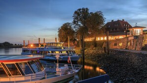 A boat docked at a riverside with a stone wall and a building with lights in the background. - Zollenspieker Fährhaus (Hamburg)