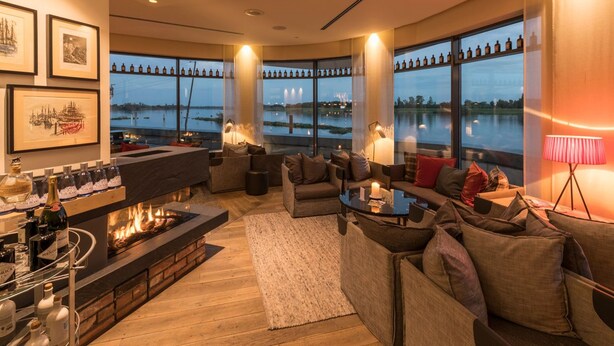 A modern living room with a fireplace, comfortable seating, a bar area with bottles, and large windows overlooking a body of water. - Zollenspieker Fährhaus (Hamburg)