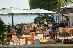 Ein Sitzbereich im Freien mit Holztischen und -stühlen, Sonnenschirmen und Blick auf einen See.