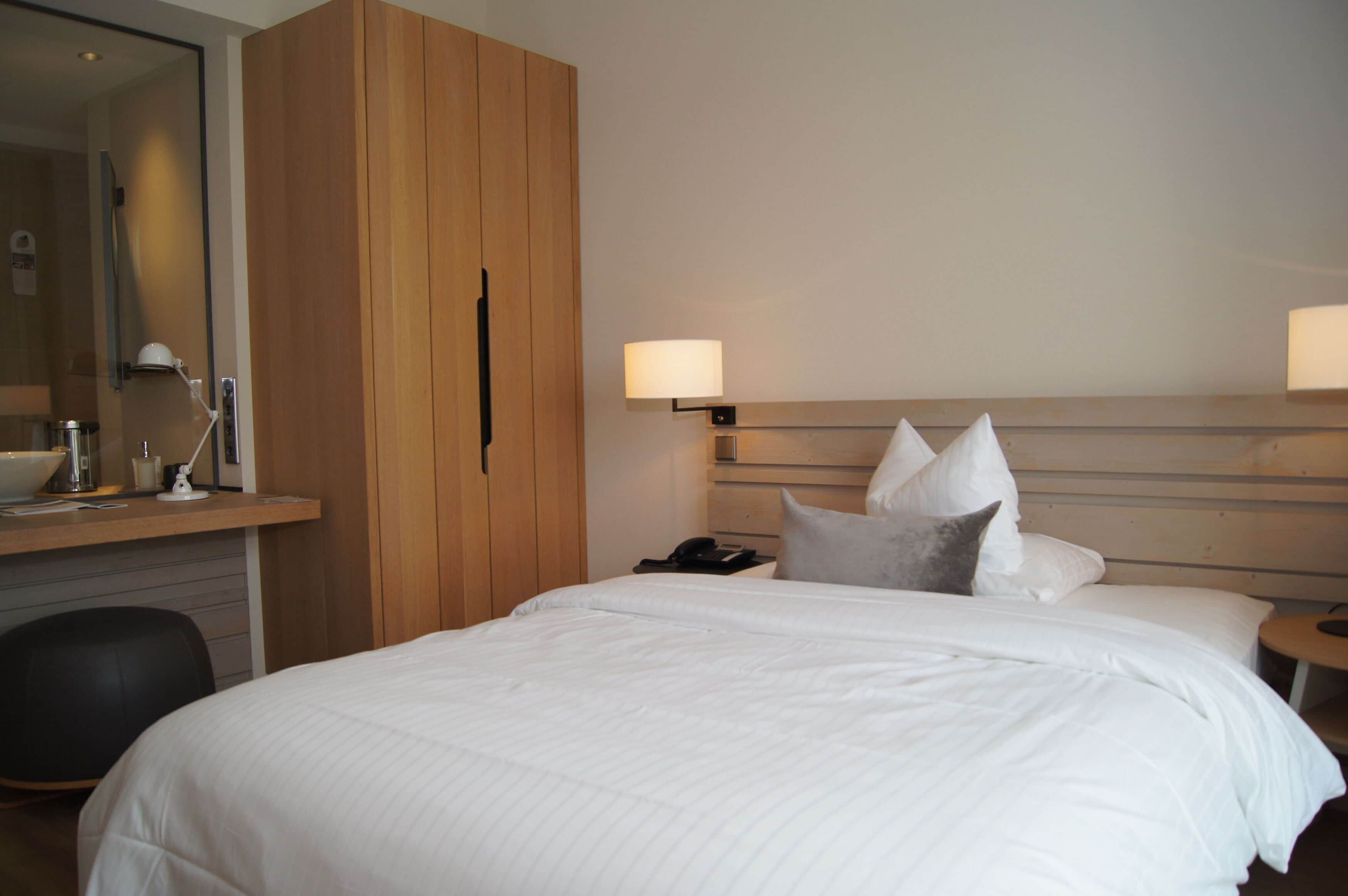 Quarto de hotel com uma cama grande, cabeceira de madeira, luminárias de cabeceira, uma escrivaninha com luminária e um espelho.