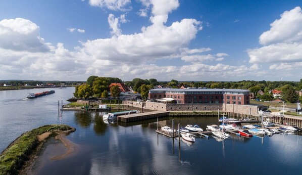 A marina with boats docked, a large building with a red roof, and a river with a cargo ship in the background.