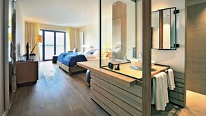 A modern hotel room with a large bed, a wooden desk, a large mirror, and a bathroom with a sink and towel rack. - Zollenspieker Fährhaus (Hamburg)