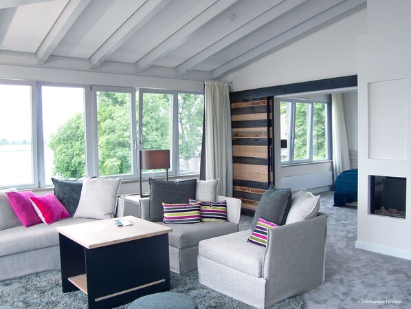 A modern living room with a sofa, armchairs, a coffee table, and a fireplace. Large windows provide natural light and views of greenery outside. - Zollenspieker Fährhaus (Hamburg)