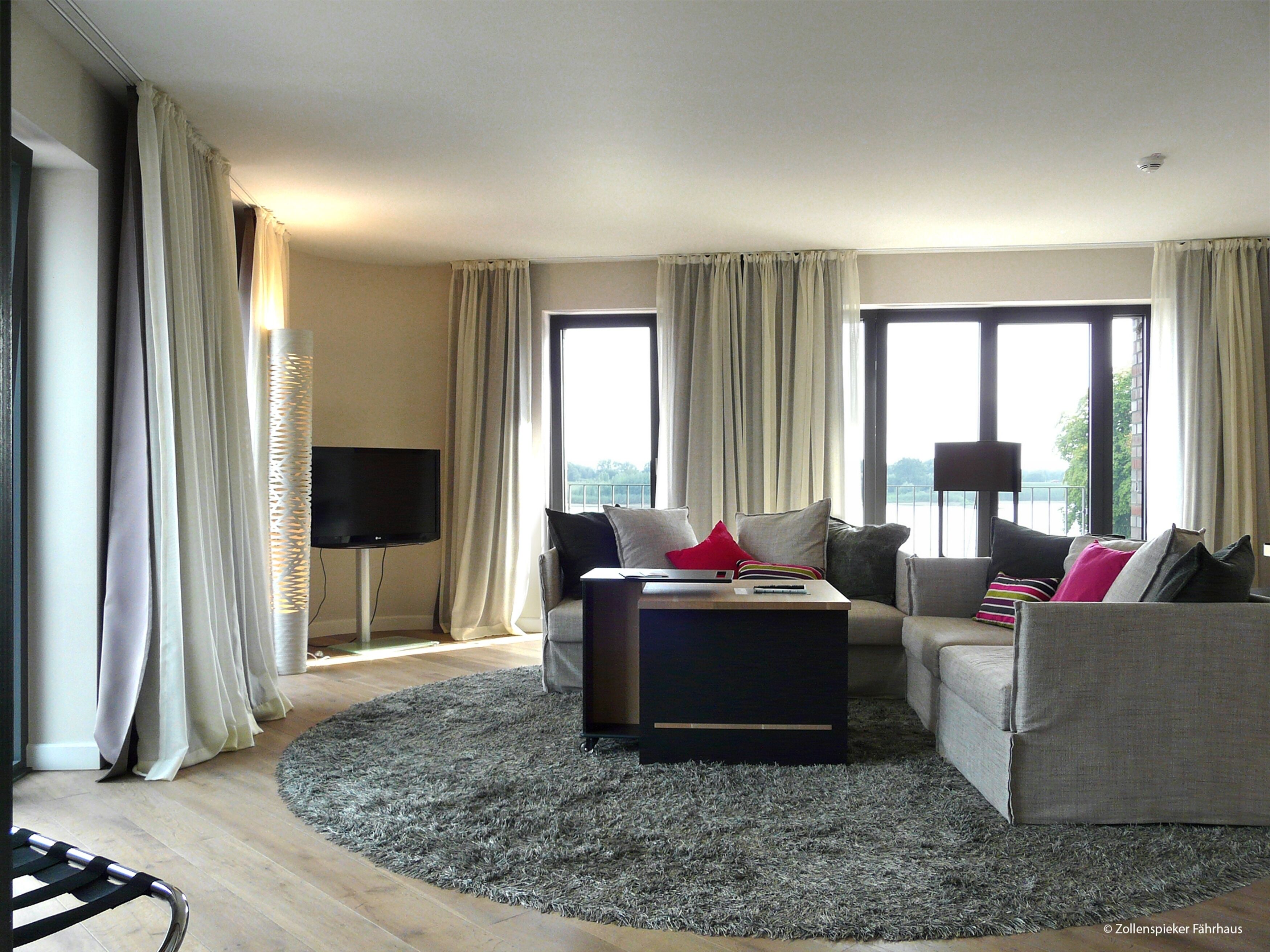A modern living room with a flat-screen TV, two sofas, a coffee table, and large windows with curtains.