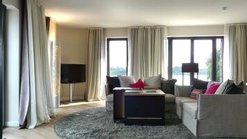 A modern living room with a flat-screen TV, two sofas, a coffee table, and large windows with curtains.