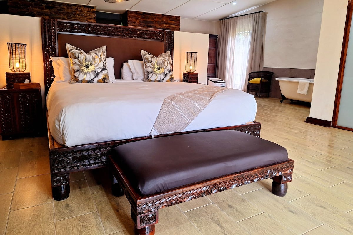 Uma cama grande com cabeceira de madeira, um banquinho e duas mesas de cabeceira com luminárias.