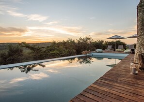 Une piscine à débordement avec une terrasse en bois, des chaises longues et des parasols, entourée d’une végétation luxuriante et offrant une vue imprenable sur le coucher de soleil.