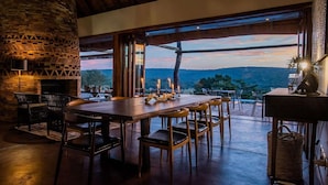 A dining area with a stone fireplace, wooden tables, and chairs, overlooking a scenic landscape. - Tala Game Reserve (Camperdown)