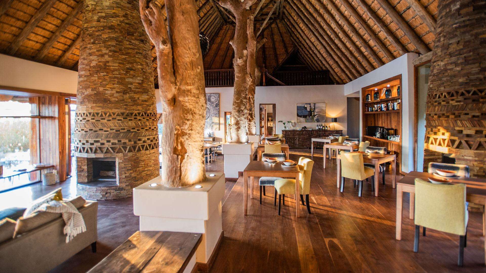 A spacious interior with a high wooden ceiling, stone walls, and a fireplace. There are multiple seating areas with tables and chairs.