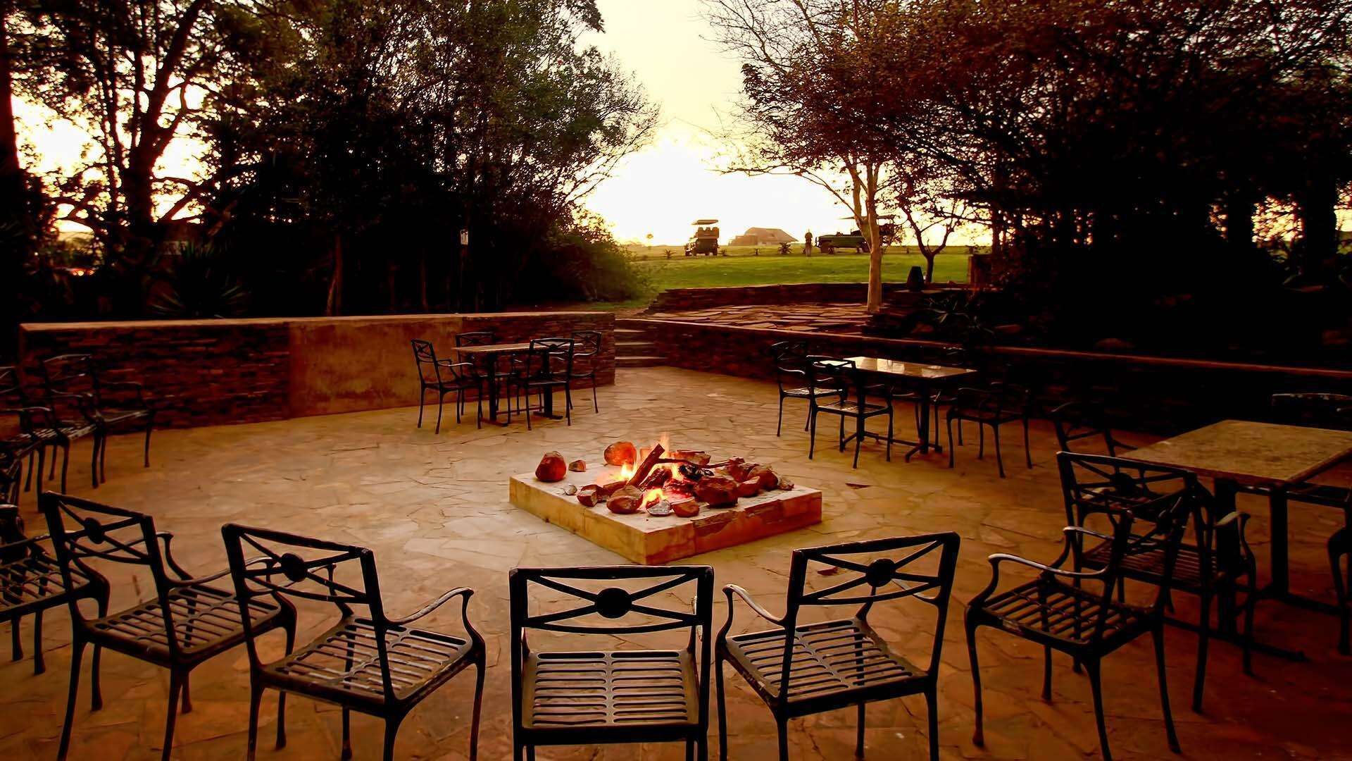 Un patio exterior con suelo de piedra, sillas de metal, un hogar y vistas a un campo.