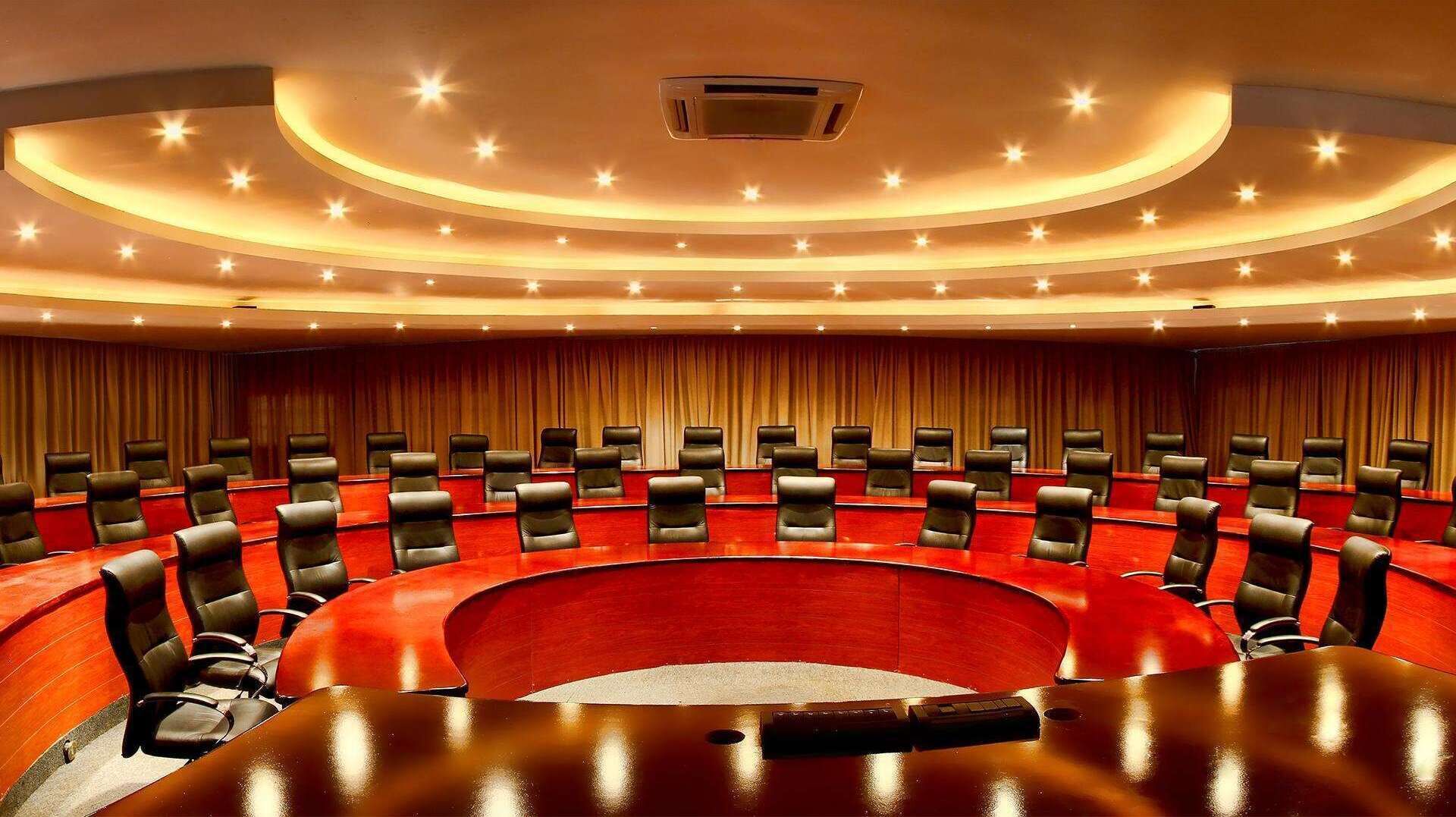 A conference room with a curved red table and black leather chairs, a tiered ceiling with recessed lighting, and a curtain backdrop.