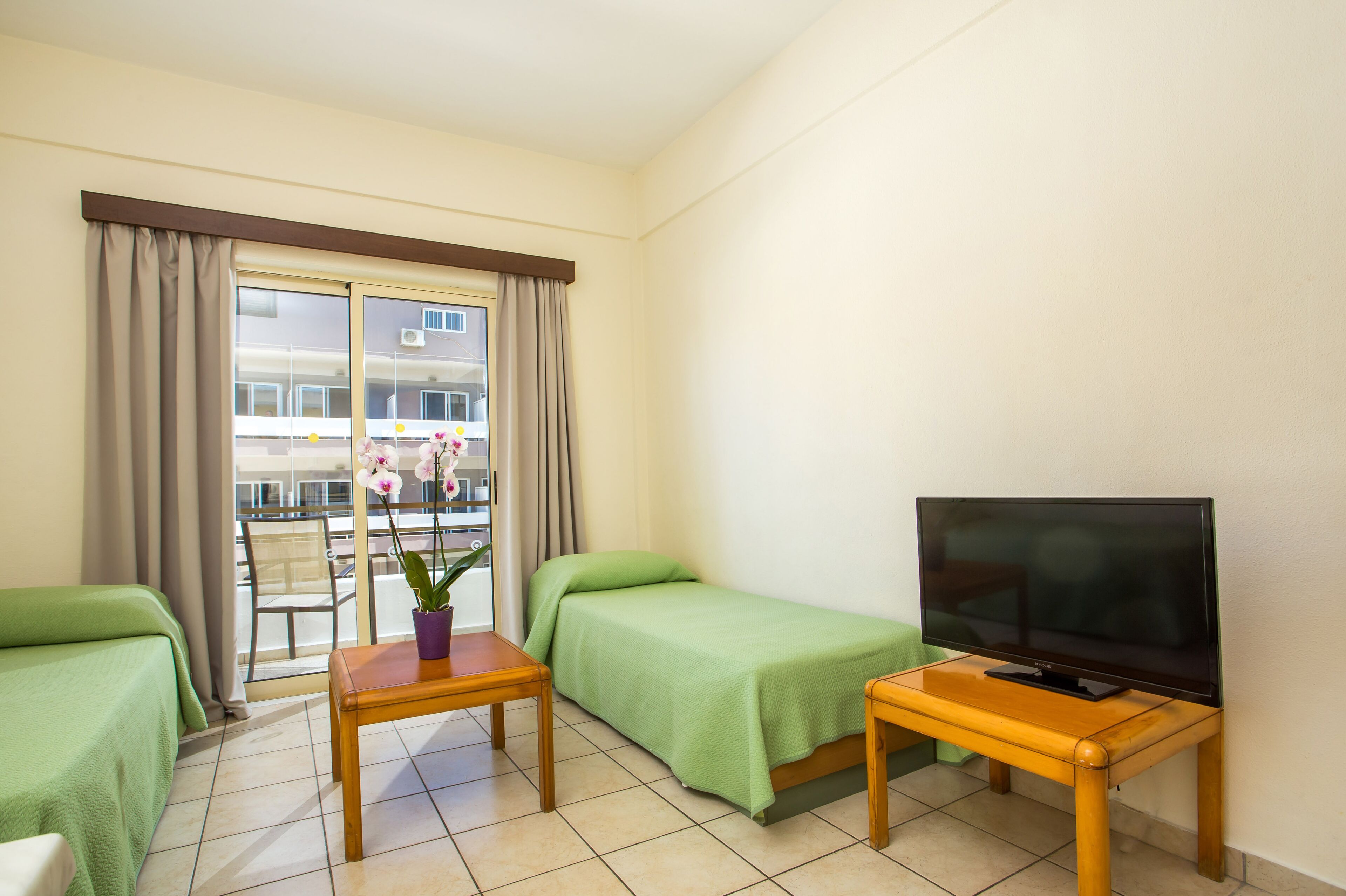 Une chambre d’hôtel avec deux lits simples, un téléviseur posé sur une table en bois et un balcon donnant sur des immeubles.