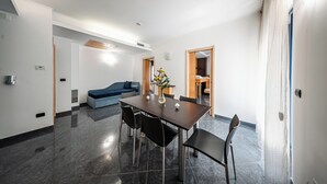A modern dining area with a rectangular table, four chairs, and a vase with flowers. There is a blue sofa and a wooden door in the background. - Blu Residence (Casarano)