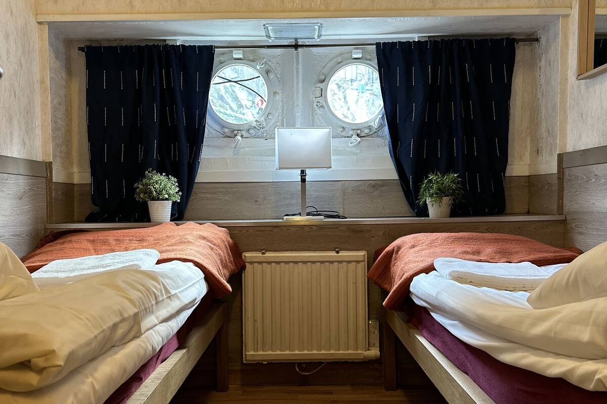 A room with two beds, a radiator, a window with curtains, a small table with a lamp, and a rug on the floor.